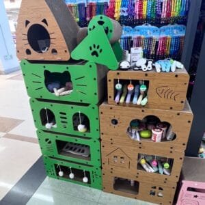 Cardboard Cat House Stackable modular cardboard cat houses in vibrant lime green and natural wood grain finishes, featuring cat-head shaped entrances, built-in scratchers, and colorful dangling play balls, displayed in a pet supply store.